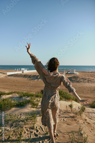 a woman in a beige dress posing standing on the seashore with her back to the camera with her arms raised high enjoying the view