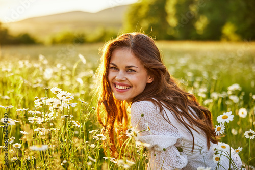 portrait of a beautiful woman in a white dress smiling at the camera sitting in a chamomile field against the backdrop of the setting sun