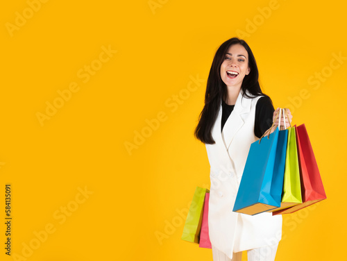 Caucasian pretty woman smiling and holding colorful shopping bags with fashion clothes and accessories inside portrait isolated on yellow background with left copy space