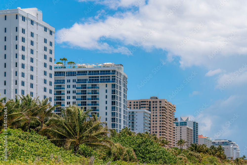 Row of multi-storey modern buildings in Miami, Florida. Apartment Hotel buildings with modern exterior near the trees and plants at the front under the giant clouds in the sky on the right.