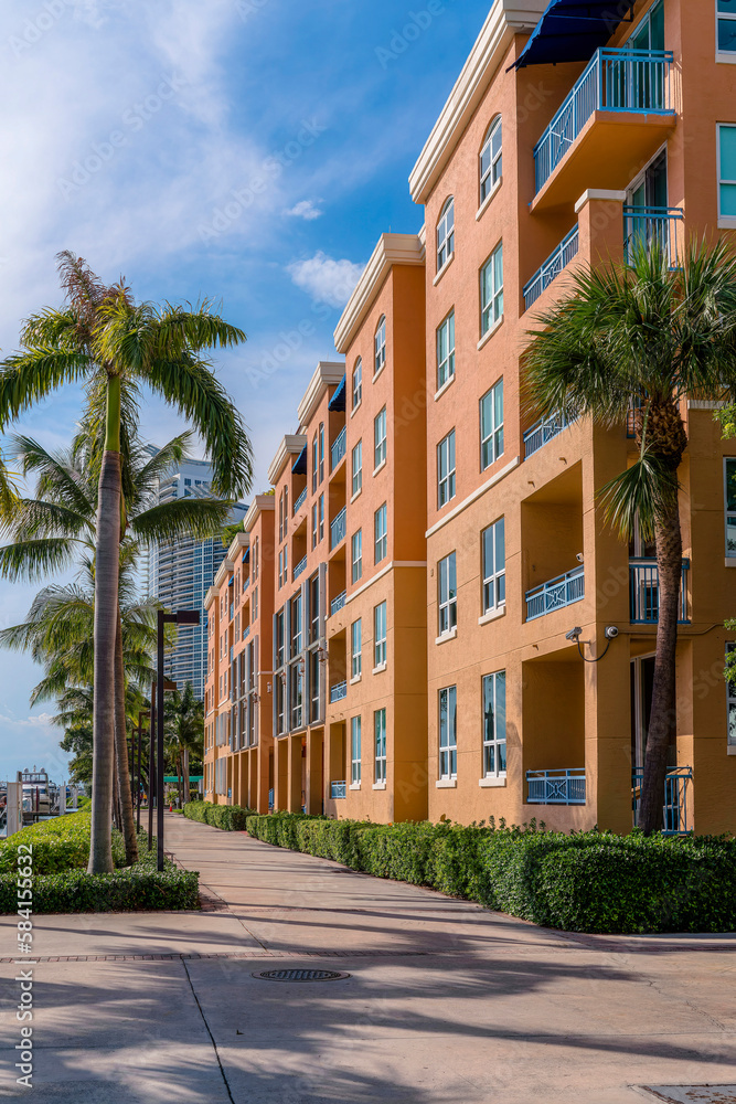 Pathway outside an orange multi-storey apartment building in Miami, Florida. Vertical shot of adjacent modern buildings with hedges and palm trees along the concrete path outdoors.