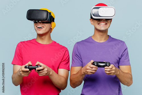 Young couple two friends men wear casual clothes together watching in vr headset pc gadget hold in hand play pc game with joystick console isolated on pastel plain light blue cyan background studio.