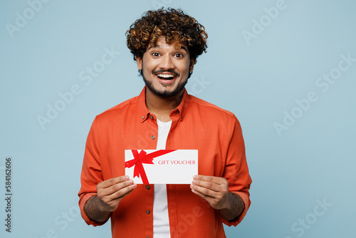 Young happy Indian man wear orange red shirt white t-shirt hold gift certificate coupon voucher card for store isolated on plain pastel light blue cyan background studio portrait. Lifestyle concept.