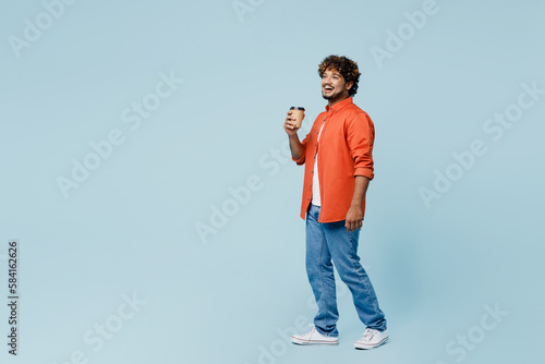 Full body young Indian man wear orange red shirt white t-shirt hold takeaway delivery craft paper brown cup coffee to go isolated on plain pastel light blue cyan background studio. Lifestyle concept.
