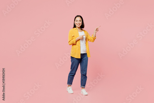 Full body young woman of Asian ethnicity wear yellow shirt white t-shirt point index finger aside indicate on workspace area copy space mock up isolated on plain pastel light pink background studio.