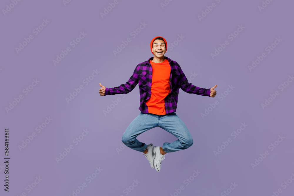 Full body satisfied young man of African American ethnicity wears casual shirt orange hat jump high spread hands show thumb up isolated on plain pastel light purple color background studio portrait.
