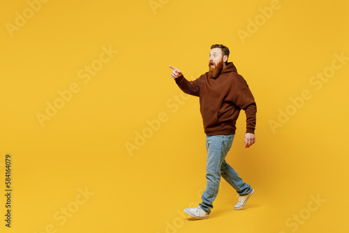 Full body smiling happy surprised shocked young redhead man wearing brown hoody casual clothes walk go point index finger aside isolated on plain yellow background studio portrait. Lifestyle concept.