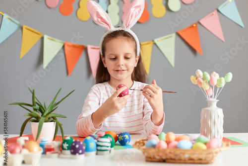 Portrait of dark haired focused little girl wearing rabbit ears sitting at table preparing for Easter coloring eggs with paint and brush against gray wall with decorations photo