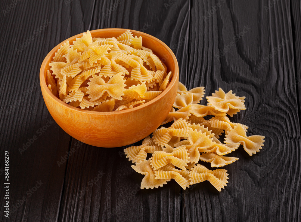 Uncooked farfalle rigate pasta in wooden bowl on black wooden background