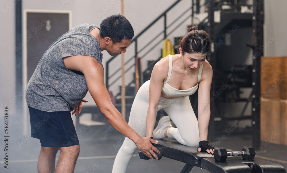 Obraz premium Asian professional muscular male personal trainer teaching female fit strong body sporty athletic fitness model in sport bra legging and gloves lifting dumbbells exercising triceps in CrossFit gym