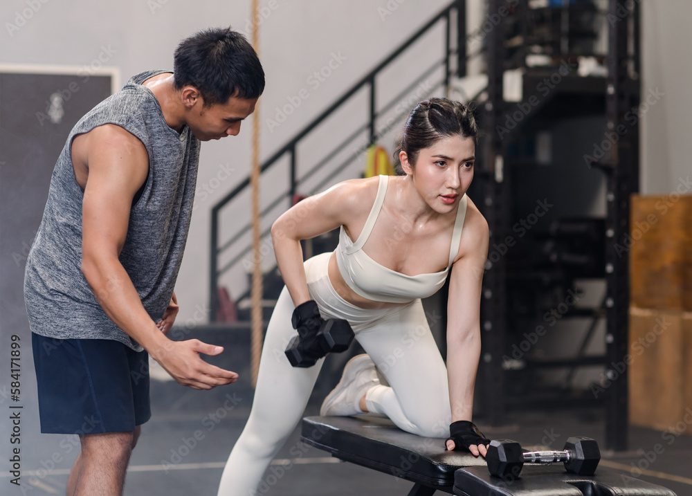 Obraz premium Asian professional muscular male personal trainer teaching female fit strong body sporty athletic fitness model in sport bra legging and gloves lifting dumbbells exercising triceps in CrossFit gym