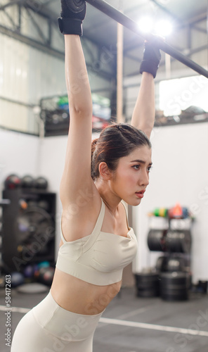 Asian young female fit strong body sporty athletic fitness model in sport bra legging and gloves hanging sweating stretching on metal gymnastic bar take break breathing after exercise training in gym