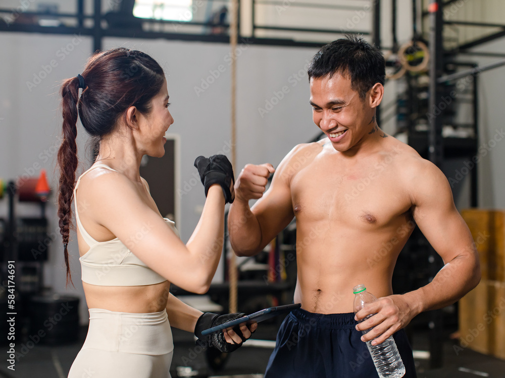 Asian professional male personal trainer take break relax rest talking with young muscular fit strong body sporty athletic sexy female fitness model holding drinking water bottle in Crossfit gym