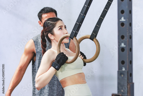 Asian young professional muscular male personal trainer teaching female fit strong body sporty athletic fitness model in sport bra legging and gloves using gymnastic hanging rings in crossfit gym