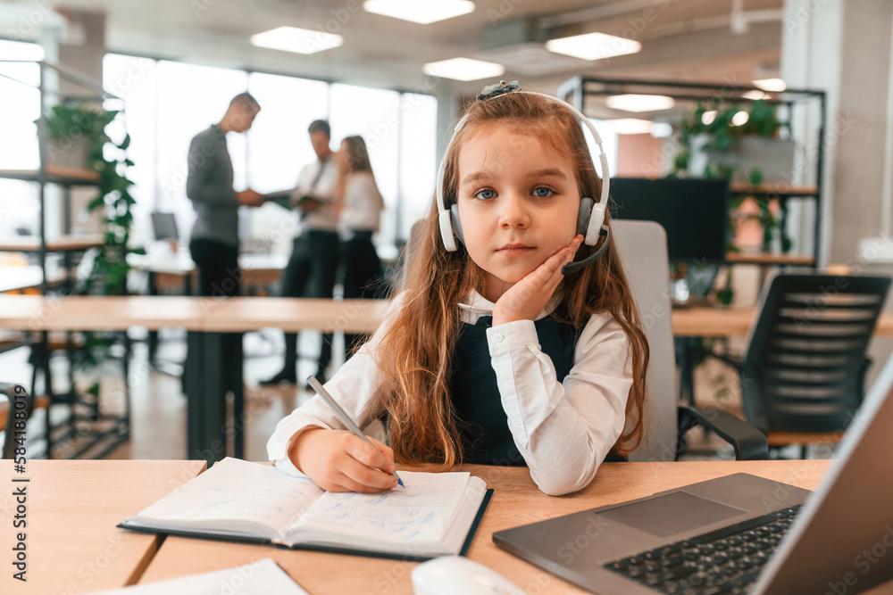Sitting by the table with laptop. Little girl is in the office where people working together Sitting by the table with laptop. Little girl is in the office where people working together