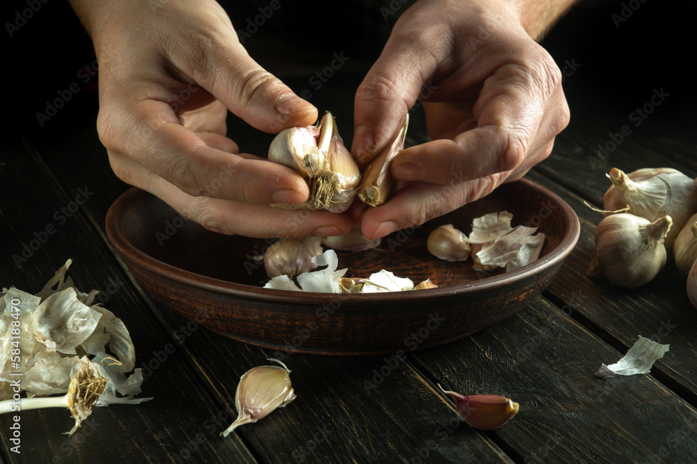 The cook hands are peeling garlic in the kitchen. The concept of cooking vegetarian food on a dark background