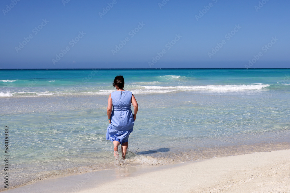 Woman in blue dress running in azure sea water. Travel and beach vacation on sandy coast