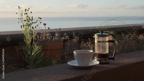 Café caliente humeante de cafetera de émbolo sobre el muro de una jardinera con luz de amanecer en una terraza con vistas al mar y mano que pone la taza en el plato