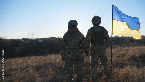 Young soldiers in camouflage uniform stands with a waving Ukraine flag at meadow against sunset. Military couple of ukrainian army lifted banner holding hands of each other at field. Dolly shot
