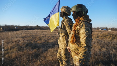 Military couple of ukrainian army lifted blue-yellow banner holding hands of each other at field. Young soldiers in camouflage uniform standing with a waving Ukraine flag at meadow. Victory at war