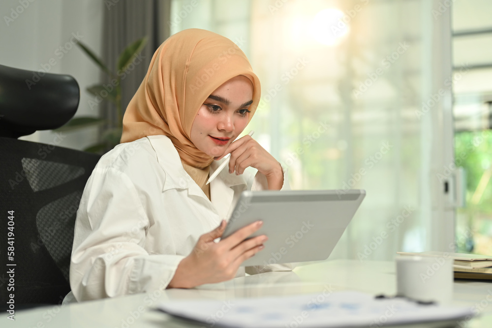 Gorgeous Muslim woman using digital tablet, remote working, searching on web at comfortable workplace