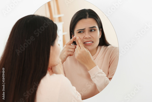 Young woman squeezing pimple near mirror indoors. Hormonal disorders