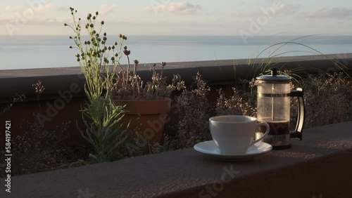 Café caliente humeante de cafetera de émbolo  con luz de amanecer en una terraza con flores y con vistas al mar con movimiento