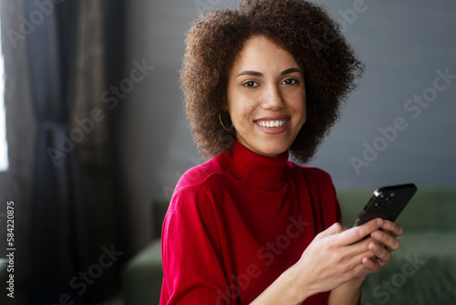 Confident African American woman in red casual attire, using mobile phone, working online, text messaging, checking social media, smiling broadly with a beautiful toothy smile, looking at camera
