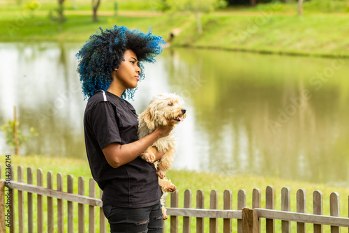 Uma jovem negra, de cabelos cacheados tingidos de azul, segurando seu cachorro no colo. photo