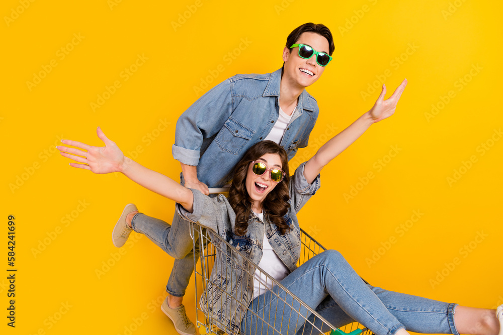 Portrait of two overjoyed excited partners push ride market trolley isolated on yellow color background