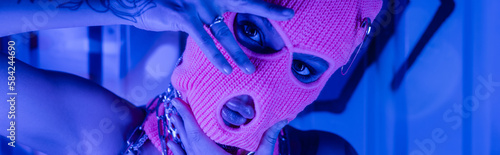 Schilderij op canvas provocative woman in balaclava posing with hands near face at wall with graffiti in blue neon lighting, banner