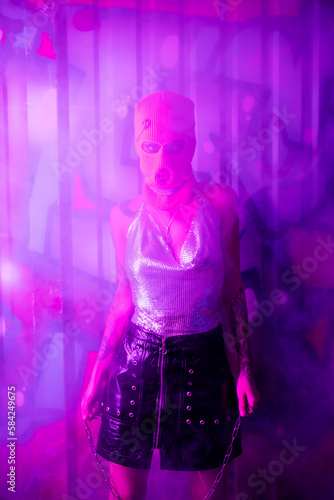 sexy woman in balaclava and silver top holding chain and looking at camera near wall with graffiti in purple light with smoke.