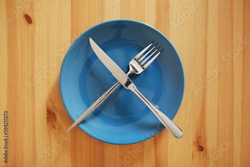 A signal "Meal break". Empty and clean blue plate with fork and knife on a wooden table as an example of table etiquette