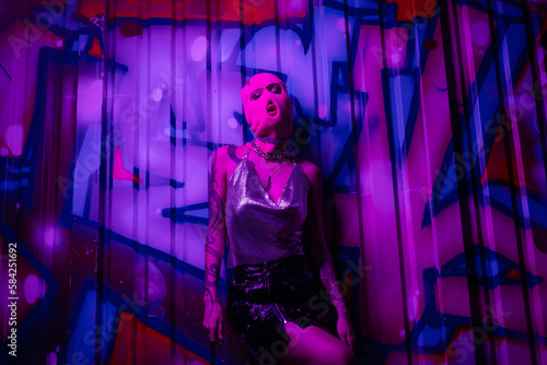 sexy woman in balaclava and silver top with black skirt posing near colorful graffiti in purple light.