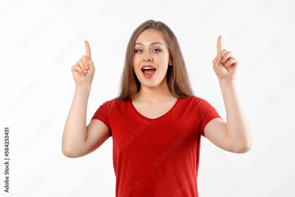 Smiling lovely girl points fingers up, shows advertisement on blank white space. Young brunette female looking happy by promotion offer, stands over white background Smiling lovely girl points fingers up, shows advertisement on blank white space. Young brunette female looking happy by promotion offer, stands over white background