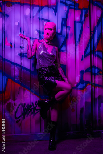 full length of stylish woman in balaclava holding silver chain and looking away near wall with graffiti in purple light.