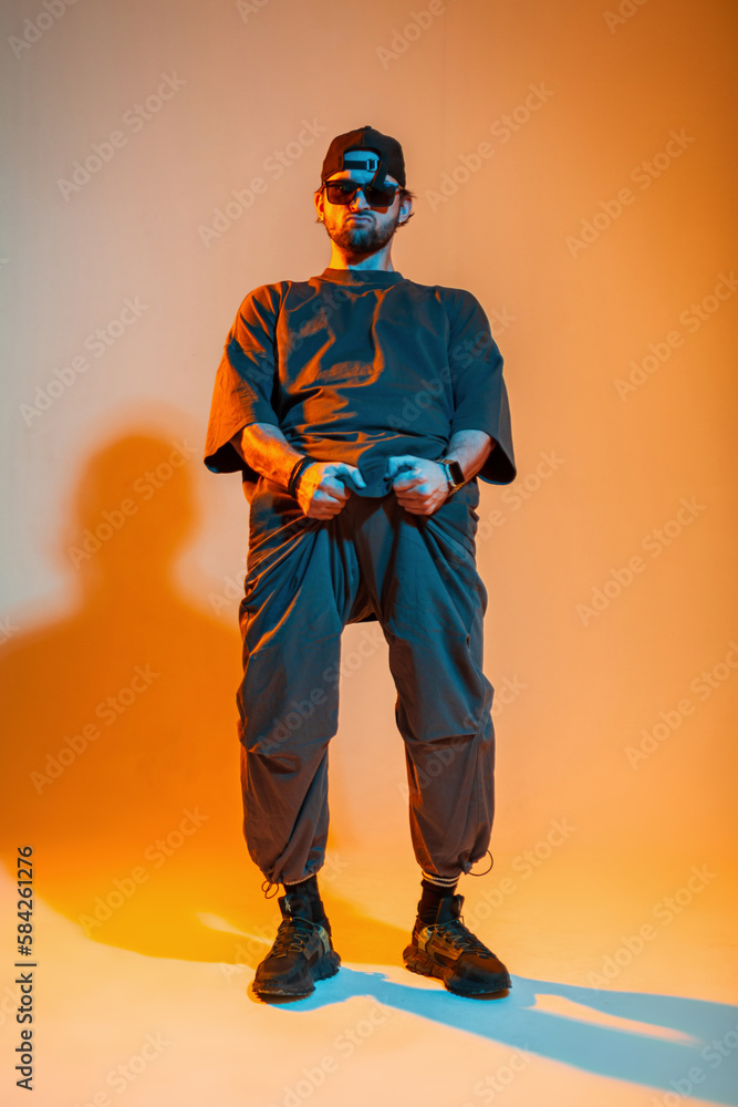 Cool stylish hipster guy with sunglasses and a cap in trendy fashion clothes holding on to his pants and poses in a creative studio with orange and neon lights Cool stylish hipster guy with sunglasses and a cap in trendy fashion clothes holding on to his pants and poses in a creative studio with orange and neon lights