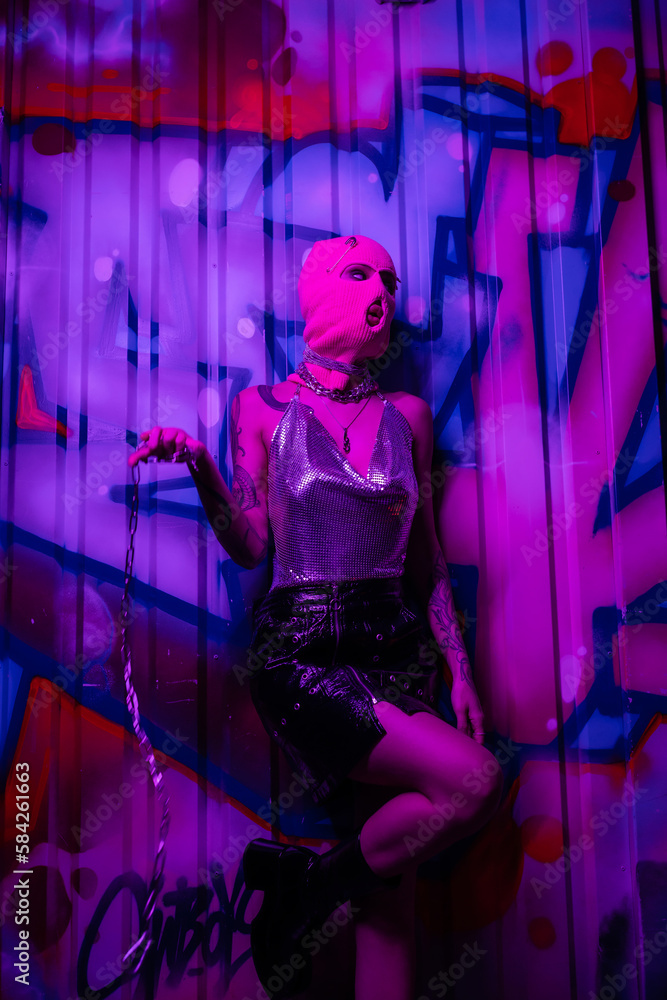 provocative woman in pink balaclava and shiny top standing with silver chain near colorful graffiti in purple neon light.