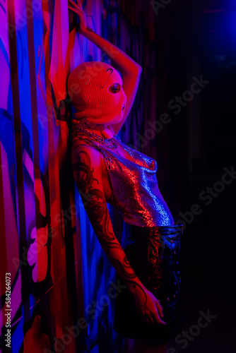 side view of tattooed woman in silver top and balaclava posing in blue and red light near wall with graffiti.