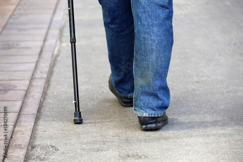 Man in jeans walking with cane on a street, legs on sidewalk. Concept of disability, limping senior adult, diseases of the spine