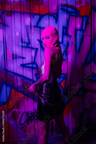 seductive woman in pink balaclava posing with silver chain and looking at camera near wall with graffiti in purple lighting.