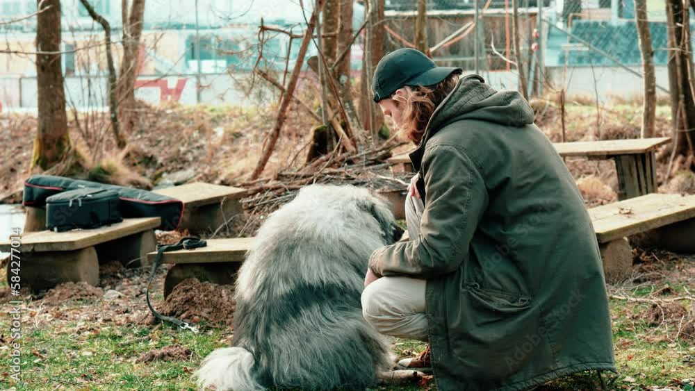here you see a young man with a black cap lovingly stroking his fluffy dog. In the background are wooden benches and a small piece of forest to see. On the benches lie bags. here you see a young man with a black cap lovingly stroking his fluffy dog. In the background are wooden benches and a small piece of forest to see. On the benches lie bags.