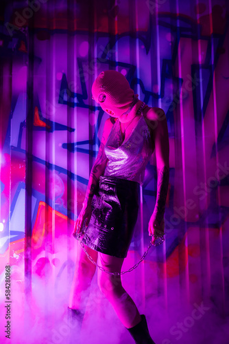 sexy woman in balaclava and shiny top with leather skirt standing with chain near colorful graffiti in purple smoke.