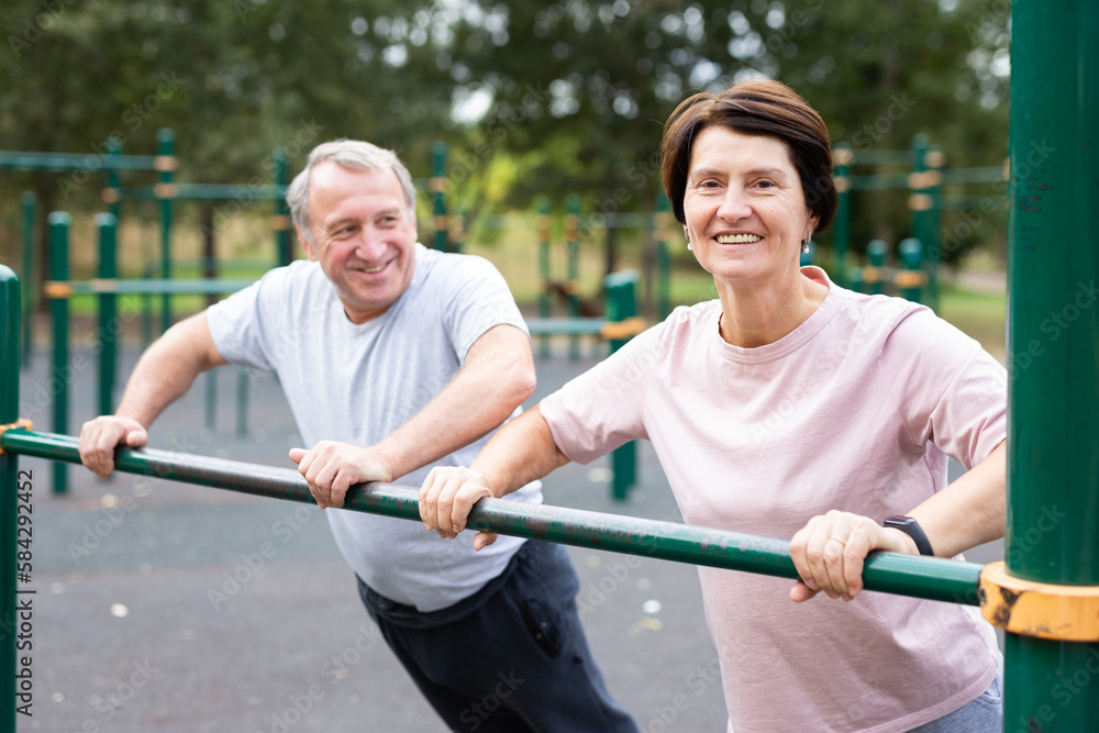 Aged man and woman doing exercises on sports bars in open-air sports area Aged man and woman doing exercises on sports bars in open-air sports area