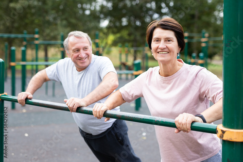 Aged man and woman doing exercises on sports bars in open-air sports area
