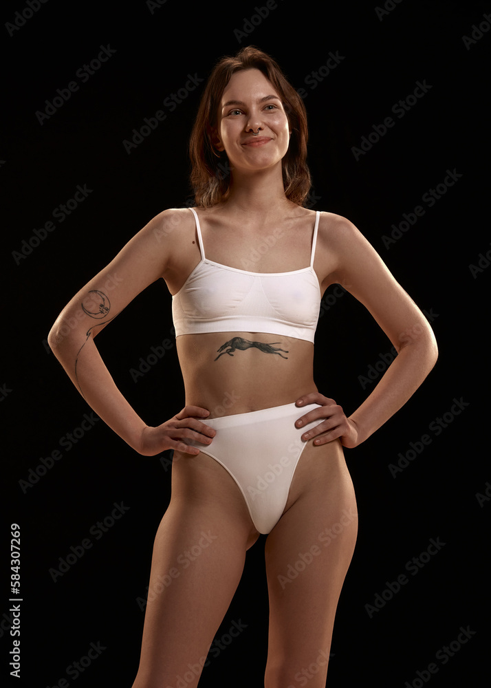 Fototapeta premium Portrait of positive, smiling , attractive young girl with tattoo and piercing posing in white cotton underwear against black studio background. Body and skin care, figure, fitness, wellness concept