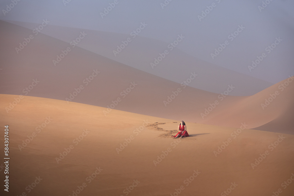 Beautiful young woman with a red dress sitting on the dunes in the desert.