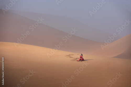Beautiful young woman with a red dress sitting on the dunes in the desert.