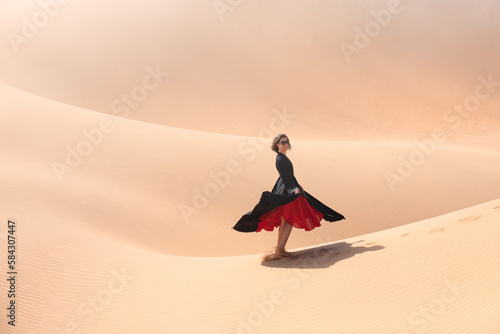 Beautiful young woman with a red dress dancing and posing on the dunes in the desert.