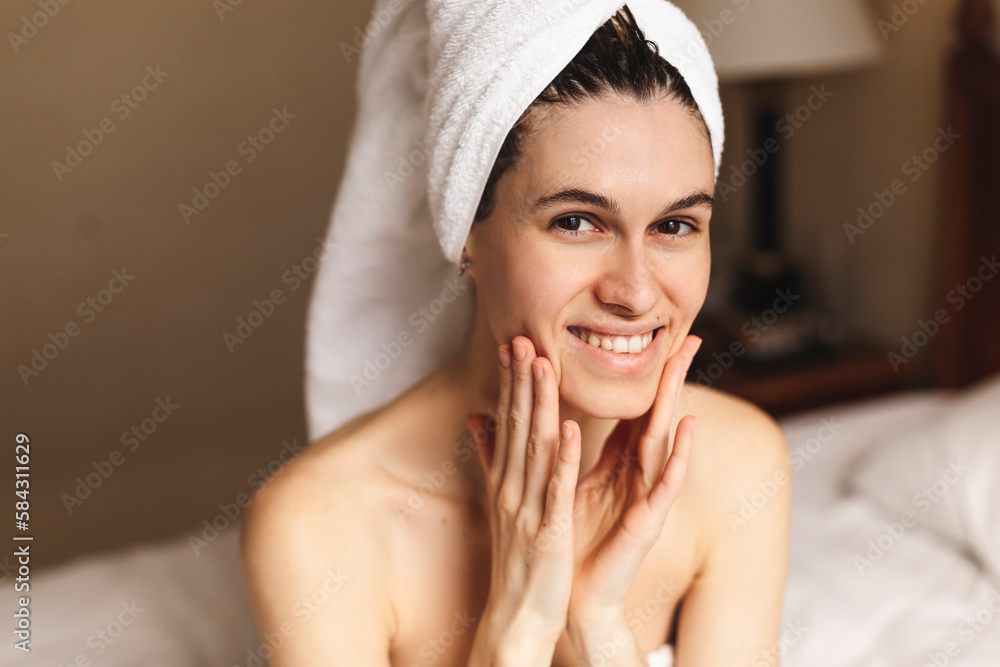 Woman in a dressing towel sits on a bed with a towel on her head smears face cream skin care beauty blogger healthy lifestyle. Spa, depilation and body care concept in hotel room. Face yoga.
